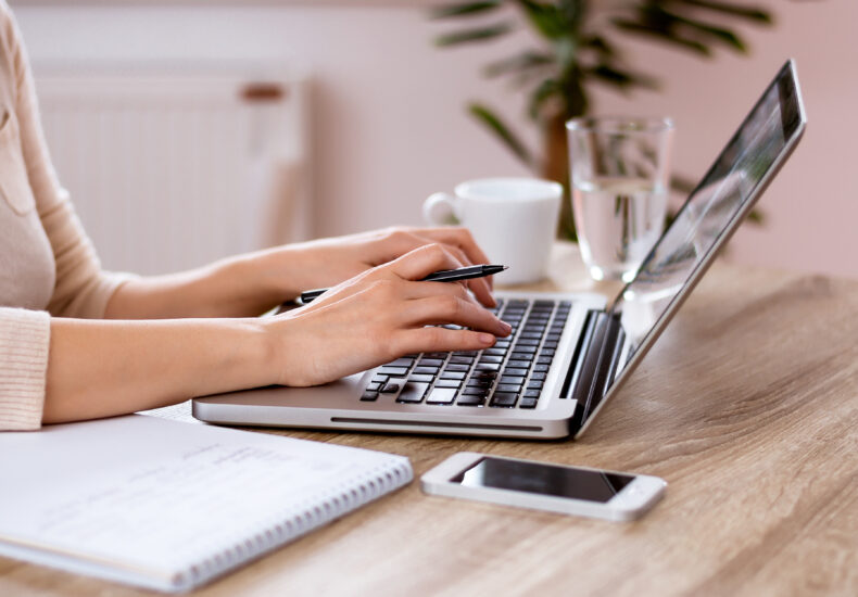 A person typing on a laptop keyboard, with a notepad and pen off to the side